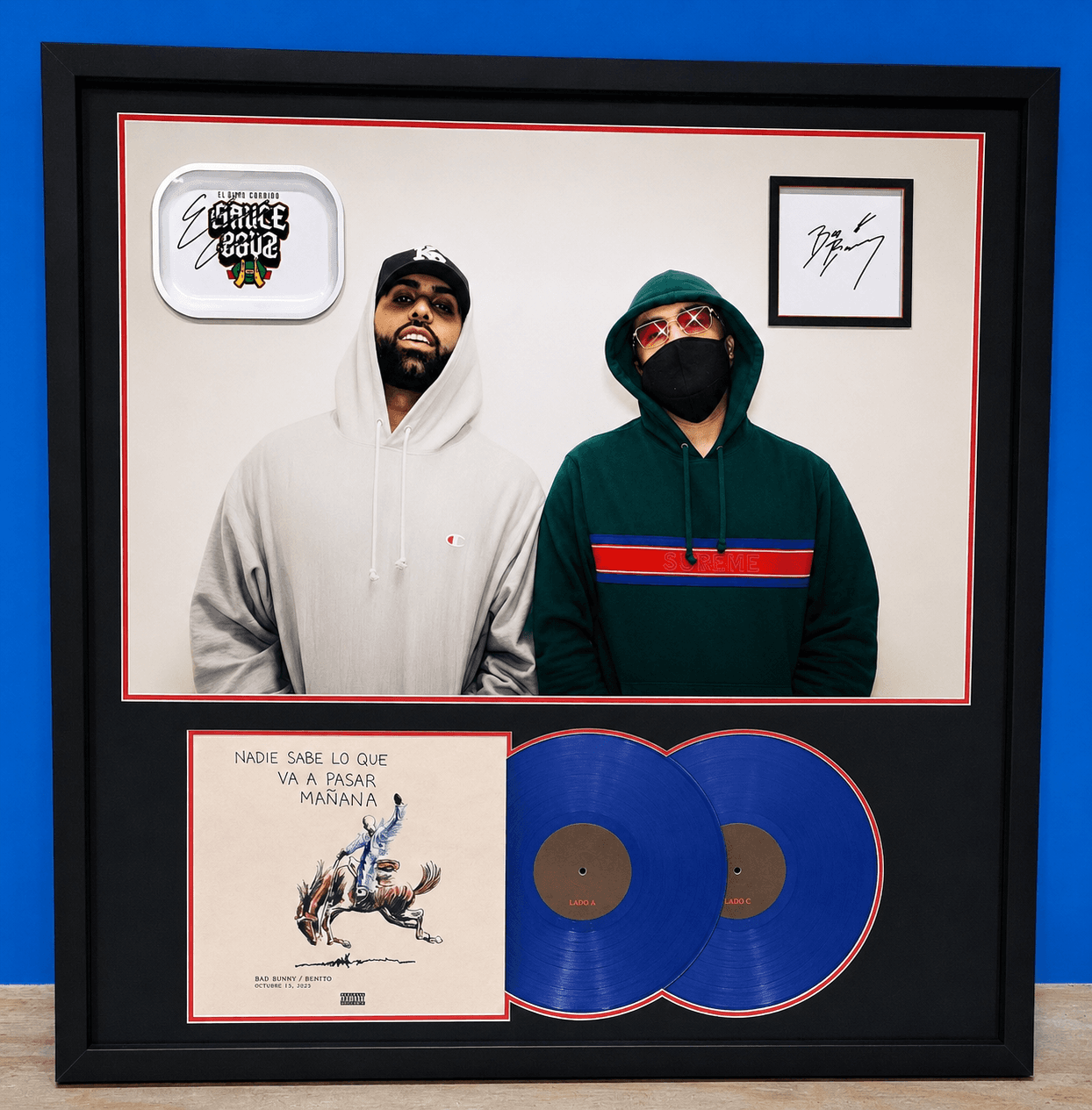 A custom multi-opening music memorabilia shadow box with a signed Bad Bunny portrait, autographed signature card, branded rolling tray, and two blue Nadie Sabe Lo Que Va A Pasar Mañana vinyl records, all matted in cream with red accent fillets and framed in a deep black moulding