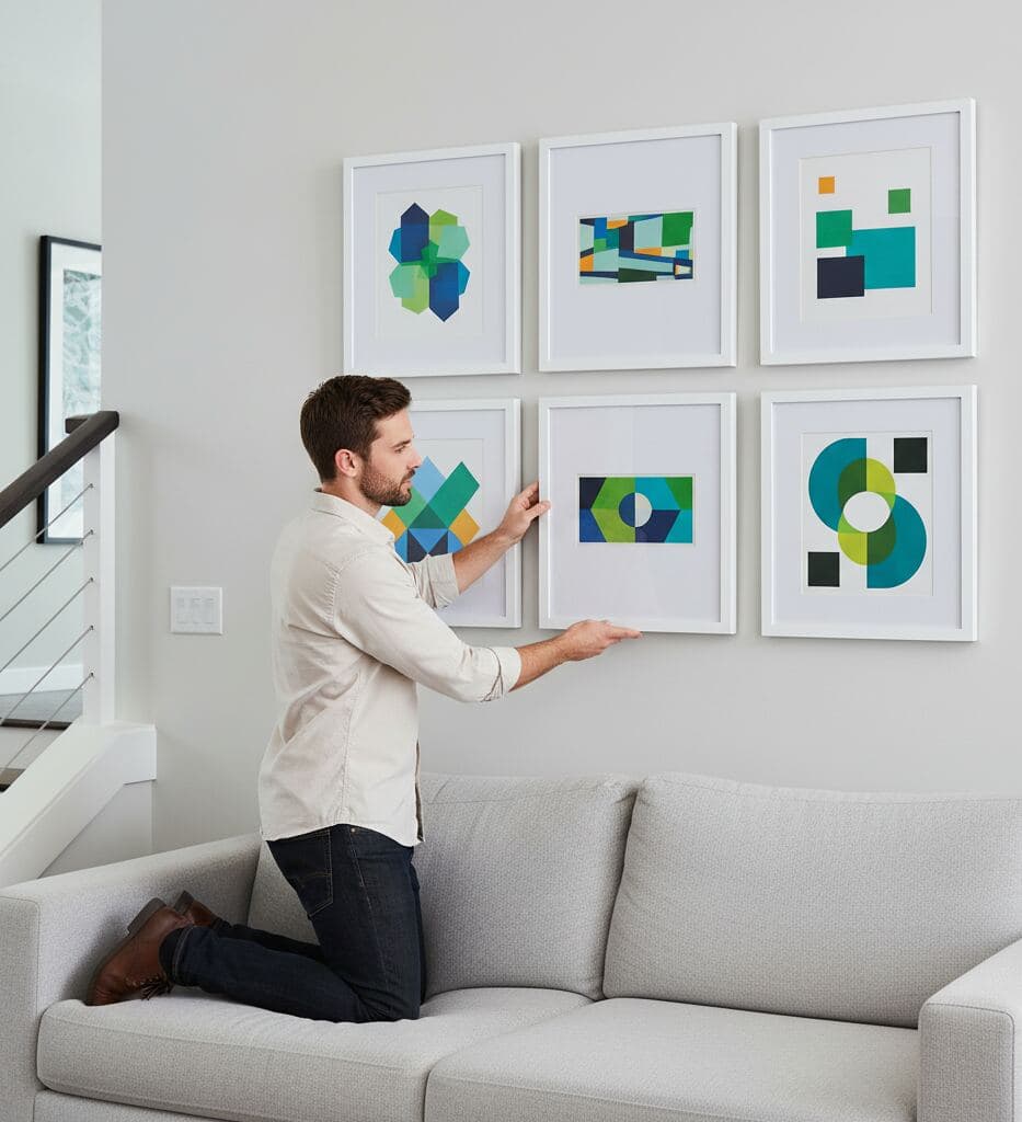Man kneels on light gray sofa adjusting grid of six white picture frames displaying geometric abstract artwork in shades of blue and green