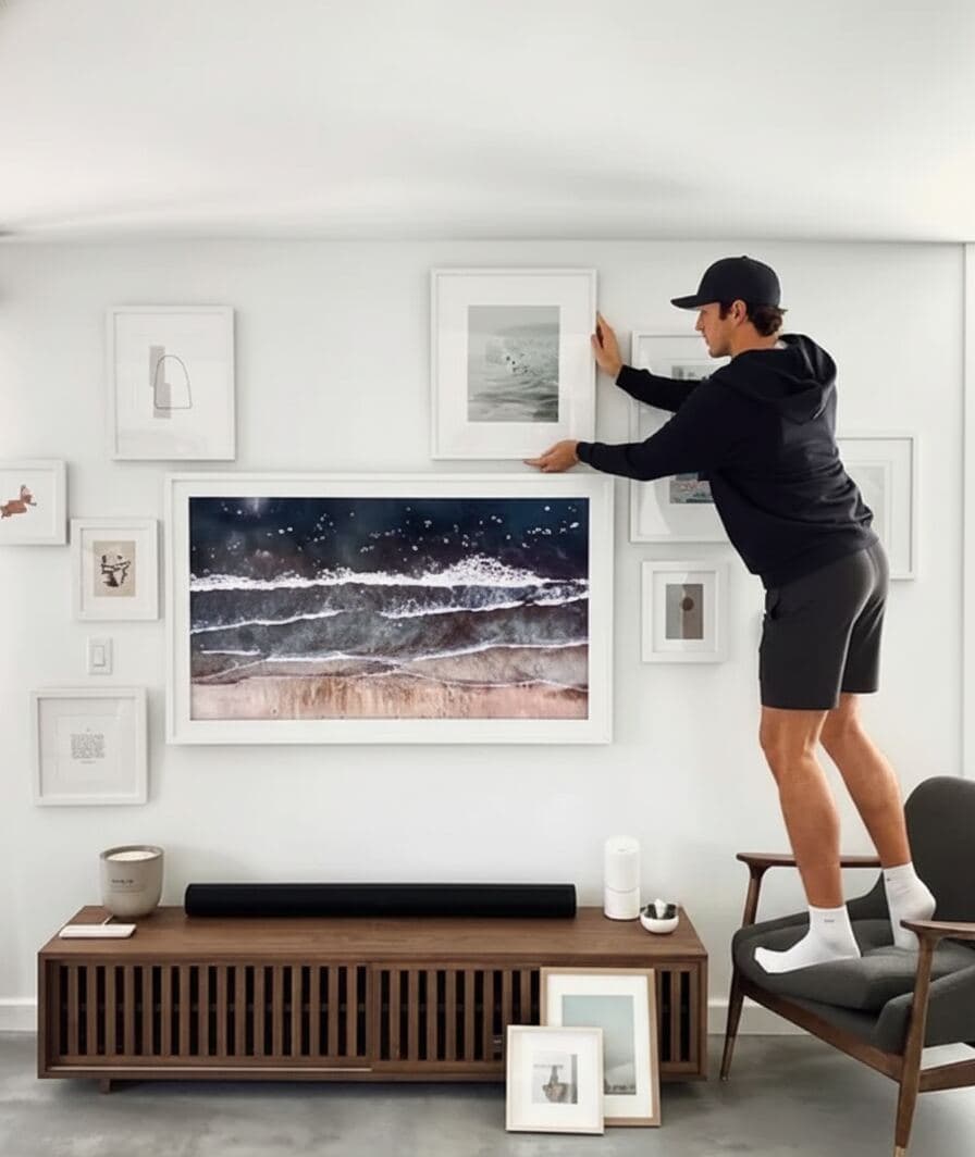 Man in dark athletic clothing stands on chair adjusting white picture frames arranged around large seascape photograph above media console