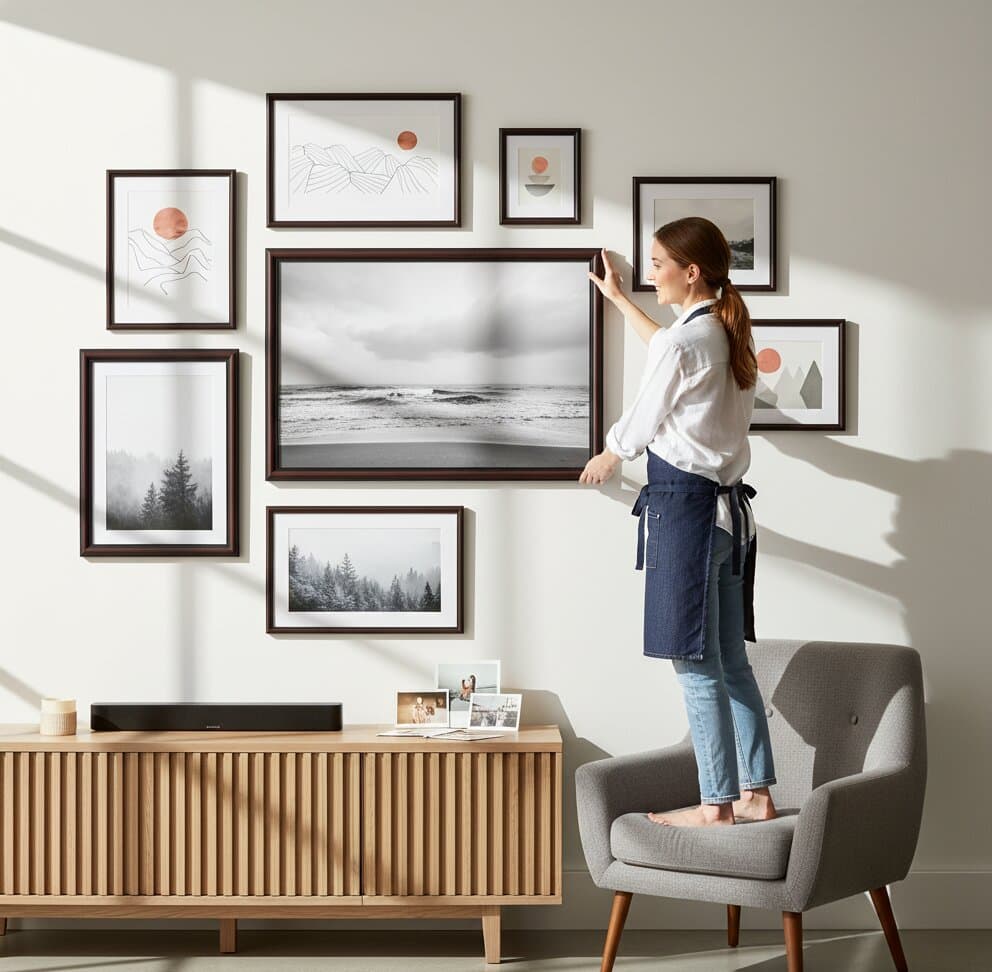 Man arranging comprehensive Walnut Picture Frame eclectic gallery wall featuring large monochromatic baby feet close-up photography flanked by vintage sepia coastal seascape palm tree botanical illustration and colorful Amalfi Coast travel poster creating balanced mixed media display above mid-century modern credenza demonstrating versatile mahogany brown finish and traditional 1.25 inch stepped moulding for mixed subject matter and family photography collections