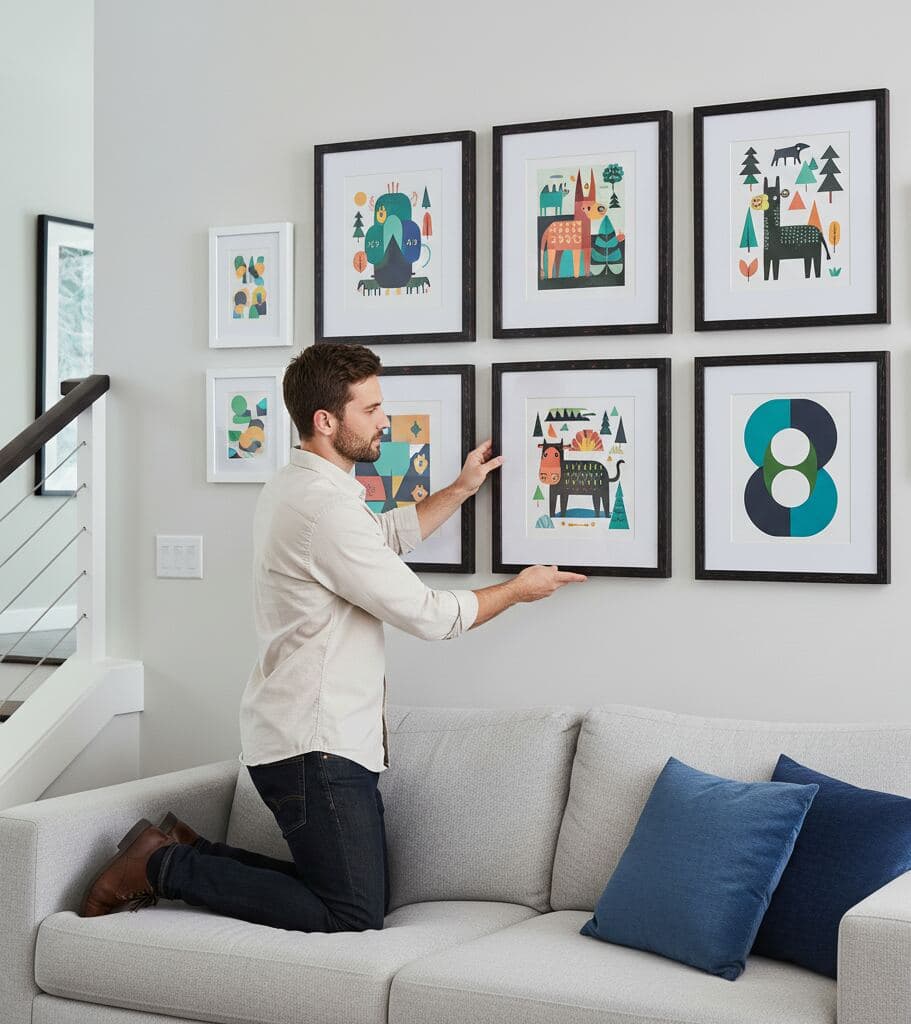 Man kneels on light gray sofa arranging gallery wall of dark wood picture frames featuring playful animal illustrations and geometric designs