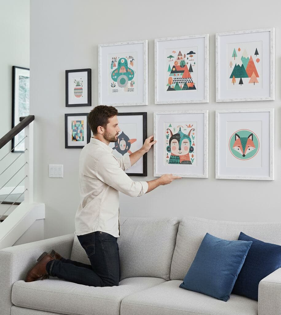 Man kneels on light gray sofa arranging wall gallery of textured white picture frames featuring geometric and animal-themed artwork