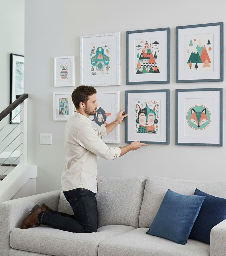 Man kneels on gray sofa arranging wall gallery of blue-washed wood picture frames featuring whimsical animal and geometric artwork