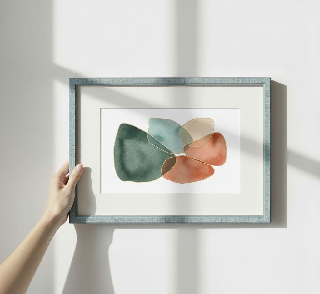 Hand holding modern silver picture frame with white mat board showcasing abstract watercolor organic shapes in teal, coral, peach, and sage green tones on minimalist wall
