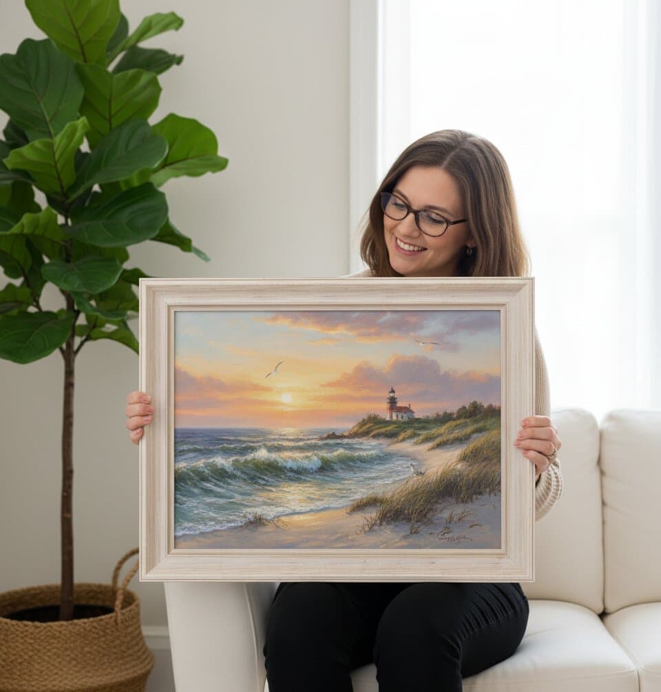 Smiling woman seated on cream sofa examines Coastal Picture Frame framed lighthouse sunset painting featuring dramatic orange pink clouds coastal lighthouse rocky shoreline and golden hour ocean reflections in substantial whitewashed wood picture frame with white matting