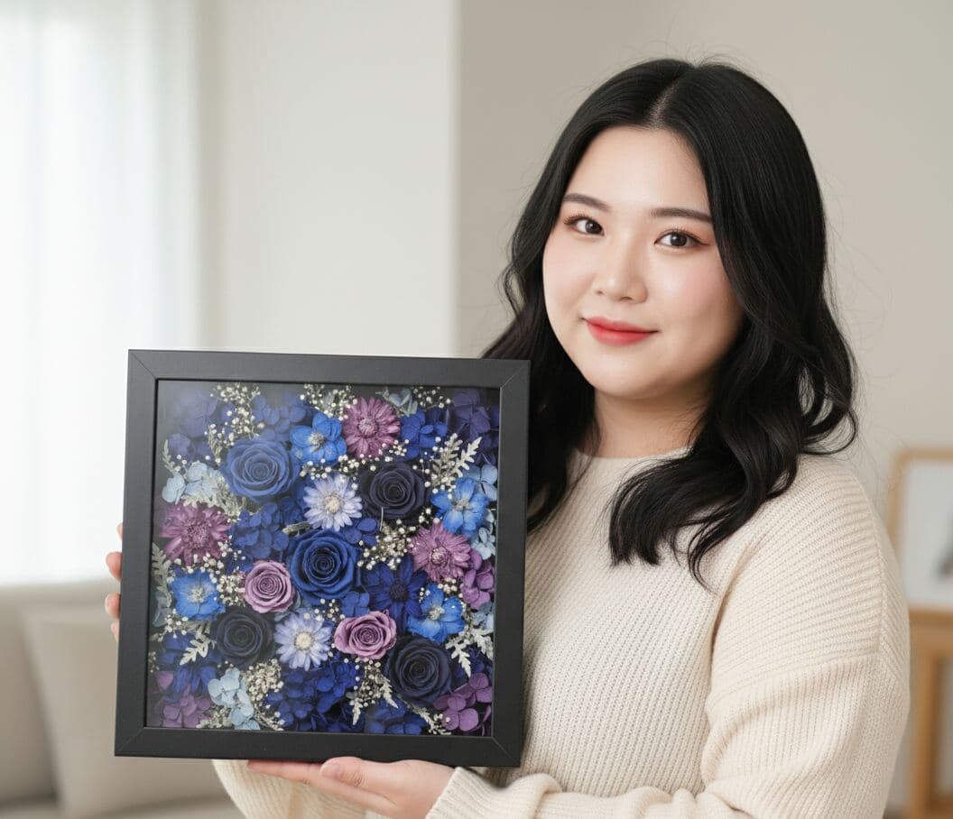 Woman holding black shadowbox picture frame with preserved blue and purple flower arrangement featuring dried roses daisies and baby's breath creating elegant floral display demonstrating custom shadow box for preserved flower art dried flower arrangements botanical keepsakes wedding bouquet preservation floral memory frames and three-dimensional pressed flower shadowbox displays
