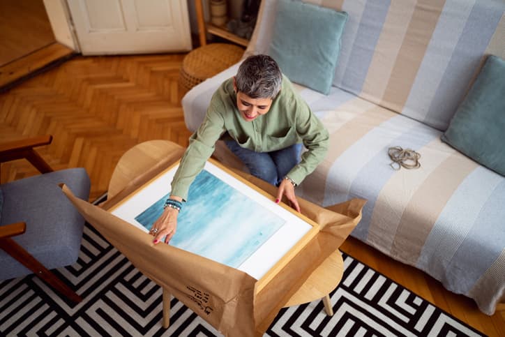 Person with short gray hair and green shirt unpacks framed blue watercolor painting on wooden coffee table in cozy living room with patterned black-and-white rug and striped sofa