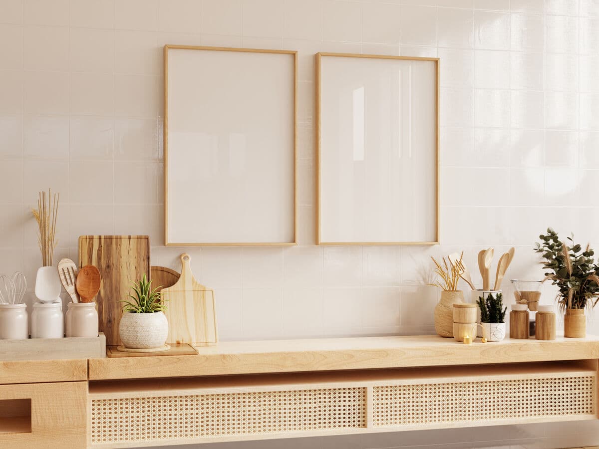 Two Natural Wood frames with blank mats hang above modern beige kitchen counter decorated with cutting boards ceramic jars wooden utensils and greenery