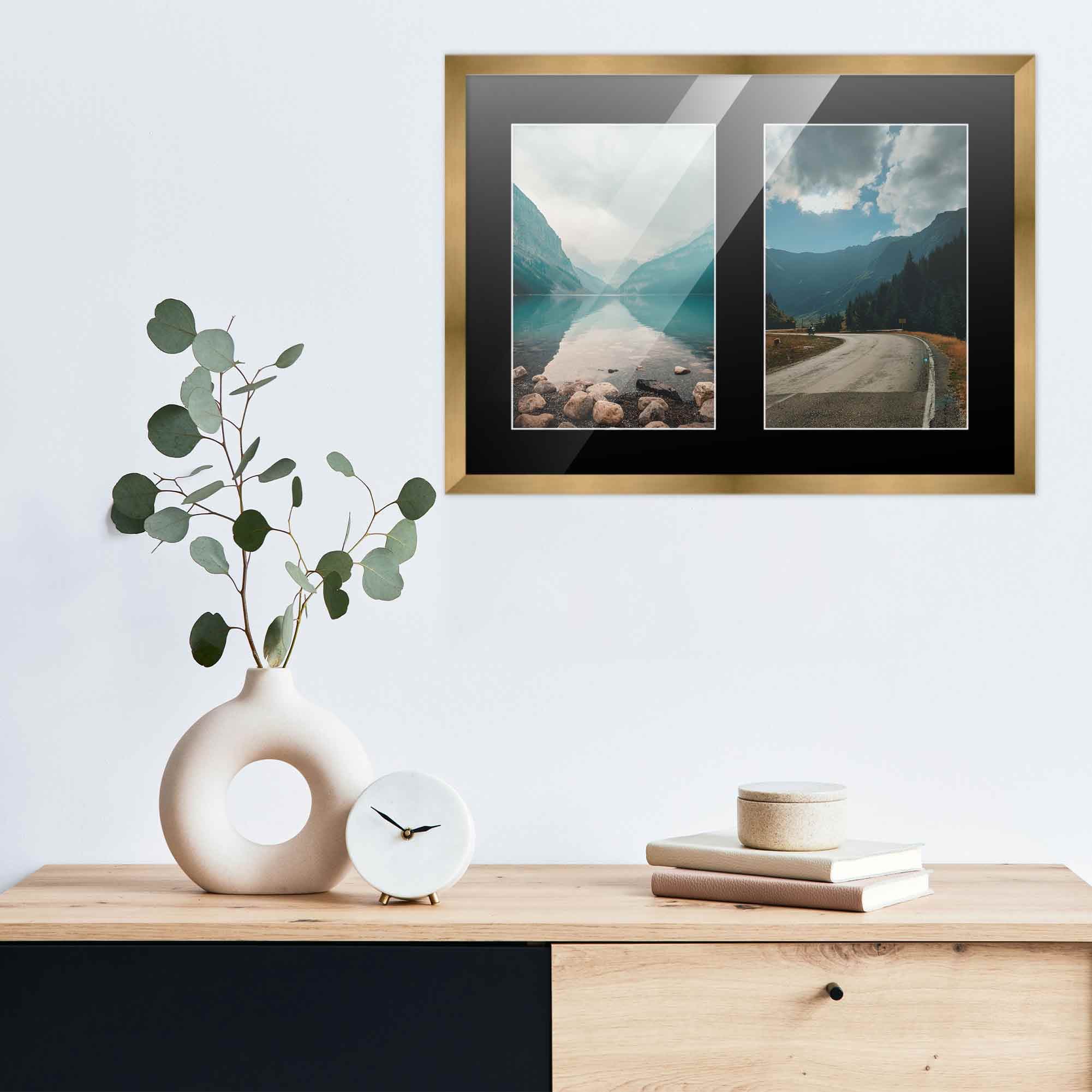 Gold double-photo frame with black mat displays two landscape photographs of lake surrounded by mountains and winding road through valley mounted above wooden sideboard with minimalist décor