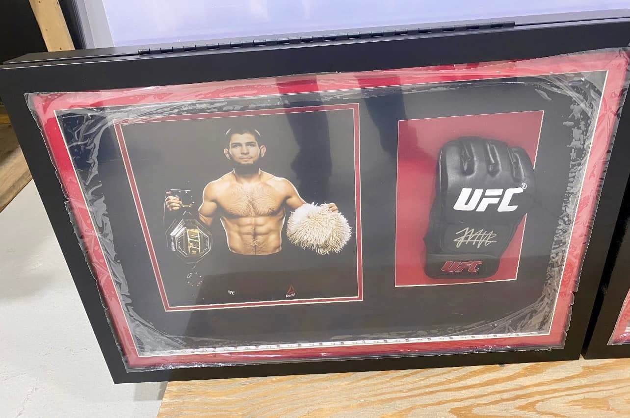 Signed Khabib Nurmagomedov UFC glove framed alongside a championship-belt print in a deep black shadow box with red double mat