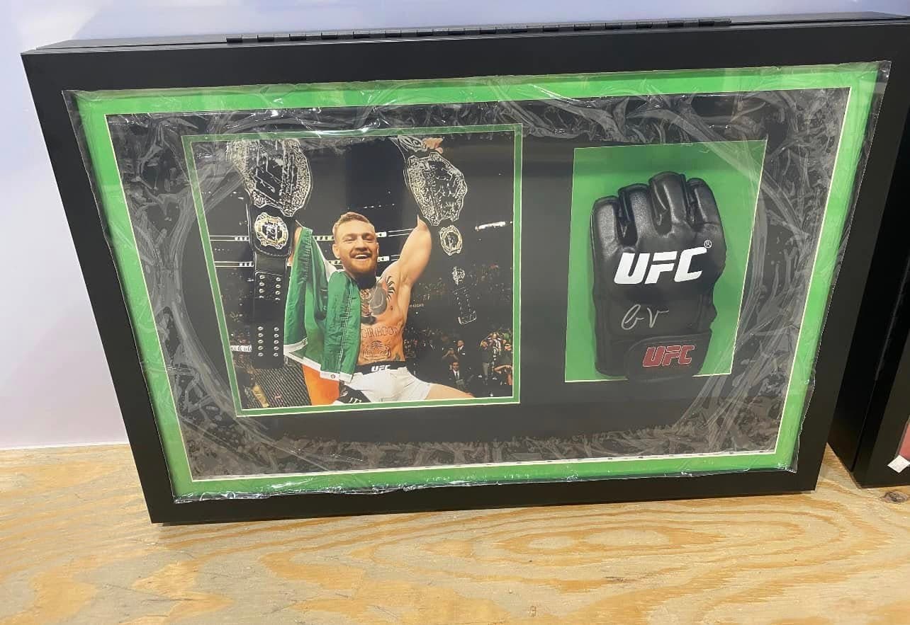 Conor McGregor two-belt champ-champ celebration shot framed alongside a signed black UFC fight glove in a wide black shadow box with Irish-green double mat and Octagon-cage background art
