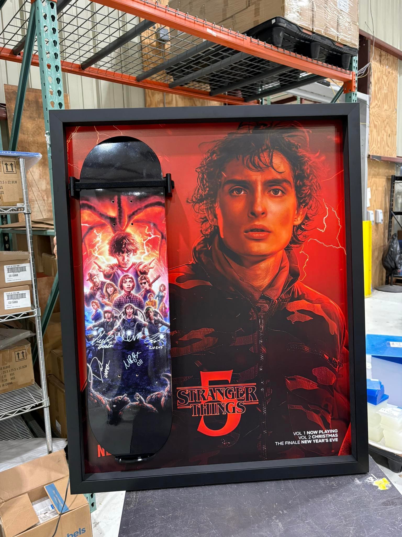 Cast-signed Stranger Things 5 skateboard deck mounted vertically against the official red Stranger Things 5 series poster inside a deep black shadow box